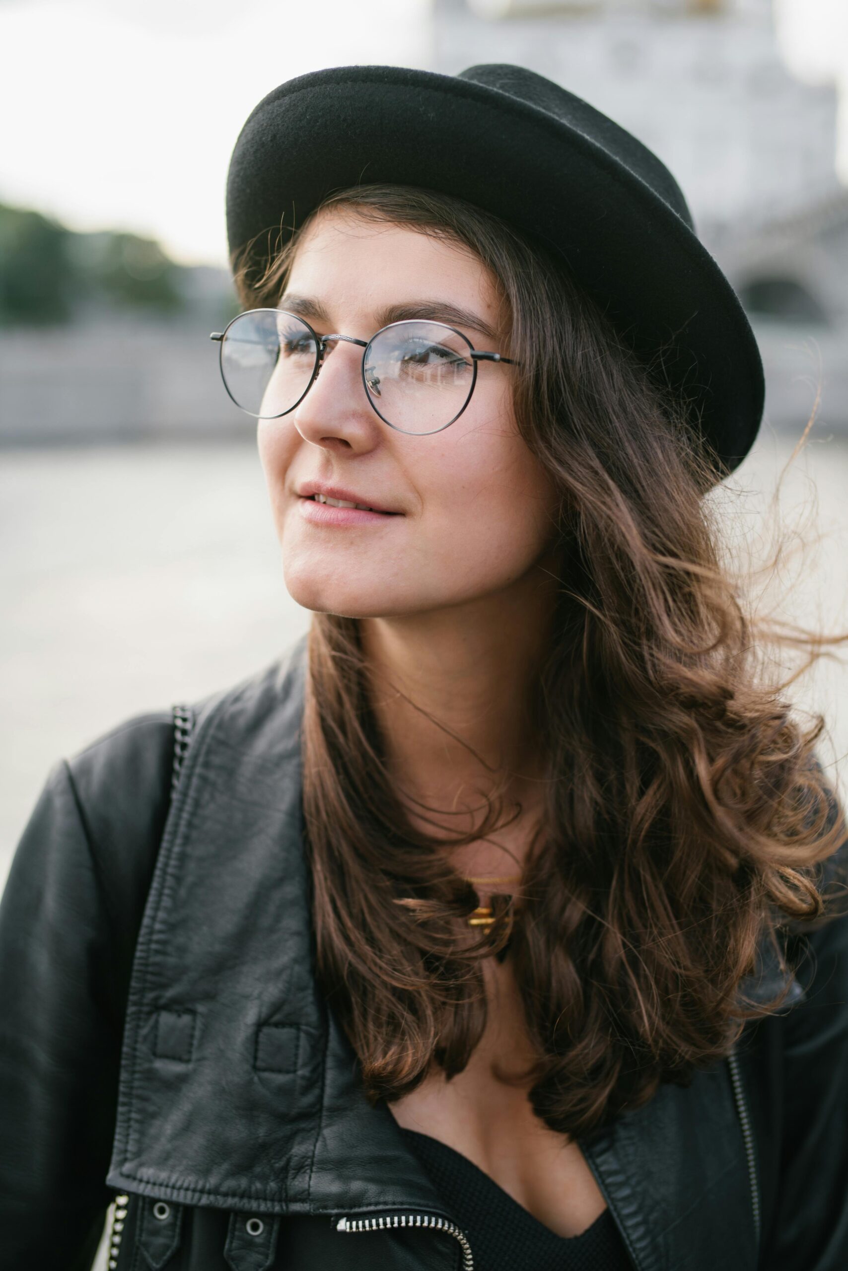 Young pleasant female in black clothes and hat and glasses thoughtfully looking away on city street